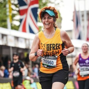Photograph of a runner participating in a race, wearing a yellow and black tank top with "Born Free Trish" text and a lion graphic, along with race bib number 70960. Background includes other runners and a large British flag, indicating a competitive event in an urban setting.