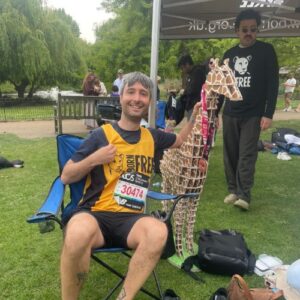 Photograph of a man sitting on a blue folding chair outdoors, wearing a yellow and black race shirt with a bib number 30474, holding a large giraffe-shaped trophy decorated with medals. Another person stands nearby under a tent, with trees and other people visible in the background, suggesting a post-race event or celebration in a park setting.