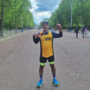 Photograph of a runner standing on a wide, tree-lined street holding up a medal and raising a fist in celebration. The runner wears a yellow and black shirt with "BORN FREE" text, bright blue shoes, and neon green shorts, with a few people walking in the background under a partly cloudy sky.