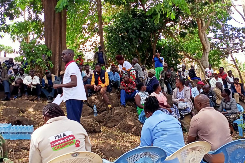 A large group of Kenyan people sit around in a circle while a main stands in the middle giving a talk