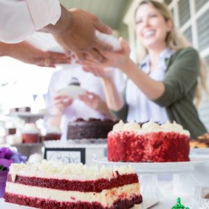 Close up of a red velvet cake, with a woman in the background handing a customer a slice of cake