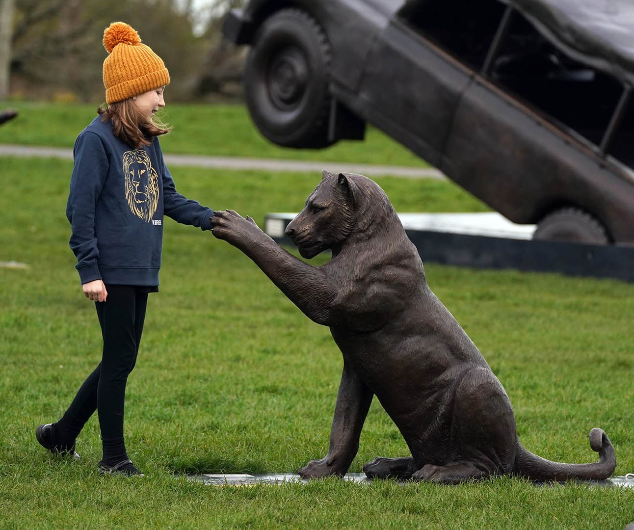 A young girl in orange bobble hat holds the outstretched paw of a bronze lion