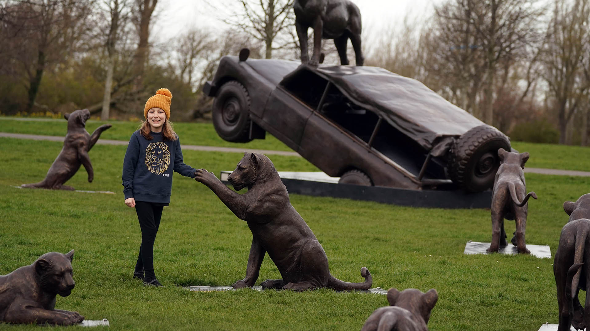 A young girl in orange bobble hat holds the outstretched paw of a bronze lion