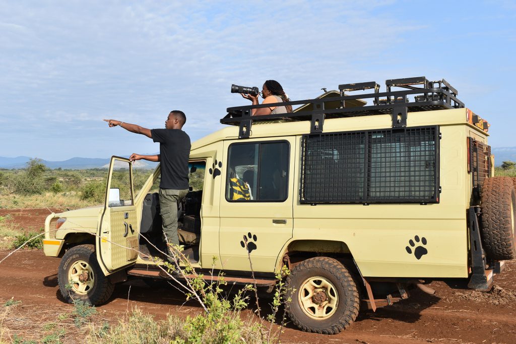 A member of Born Free's Pride of Meru team leans out of a truck pointing at something in the distance, a photographer points a camera in the same direction