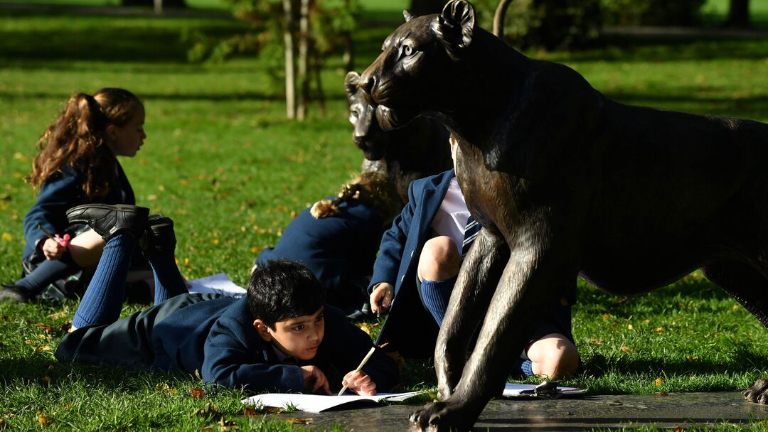 School children in uniform sit on the ground next to a bronze lion sculpture, with open books that they are drawing in