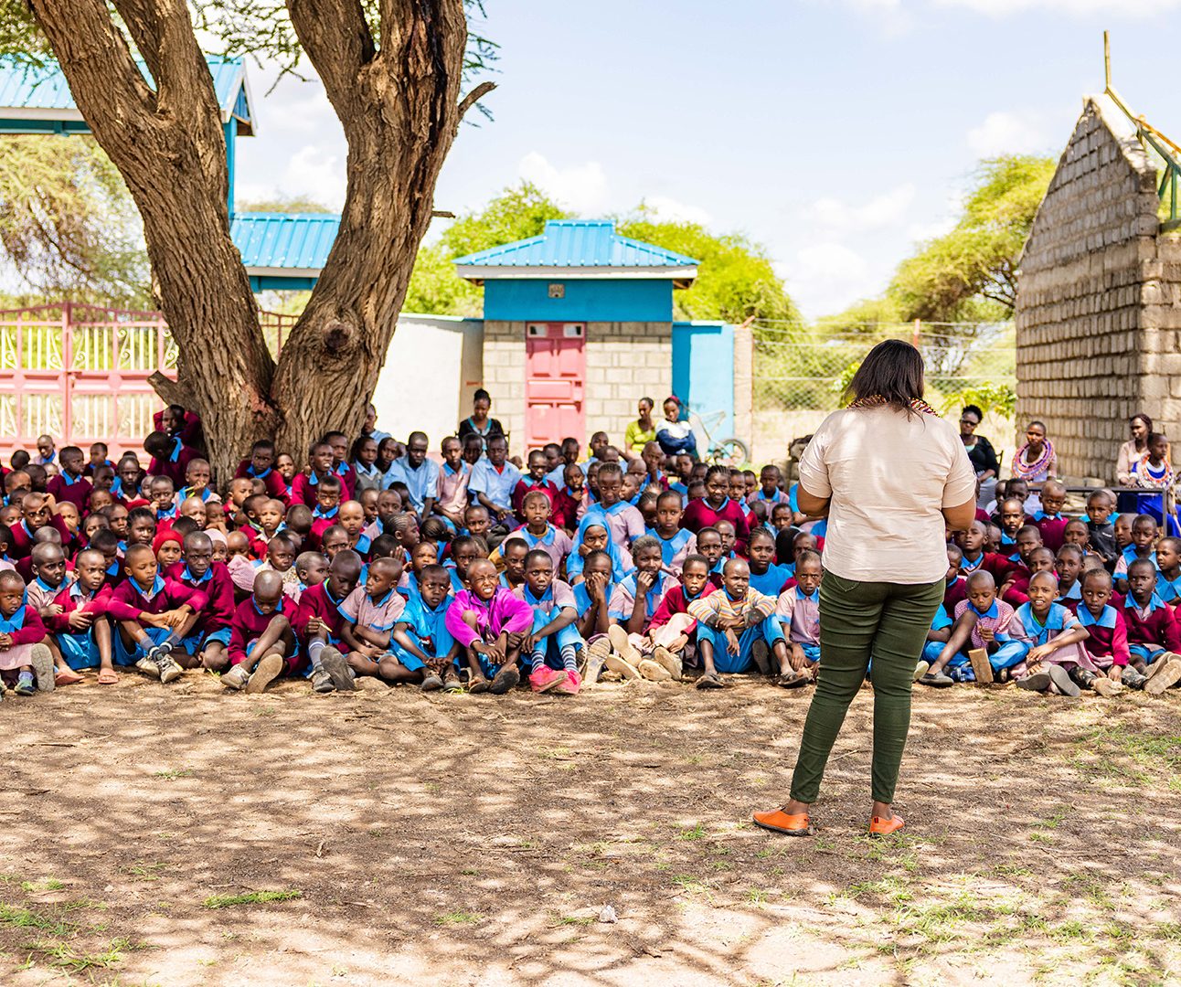 A woman stands with her back to the camera, facing a crowd of dozens of Kenyan school children sitting on the ground