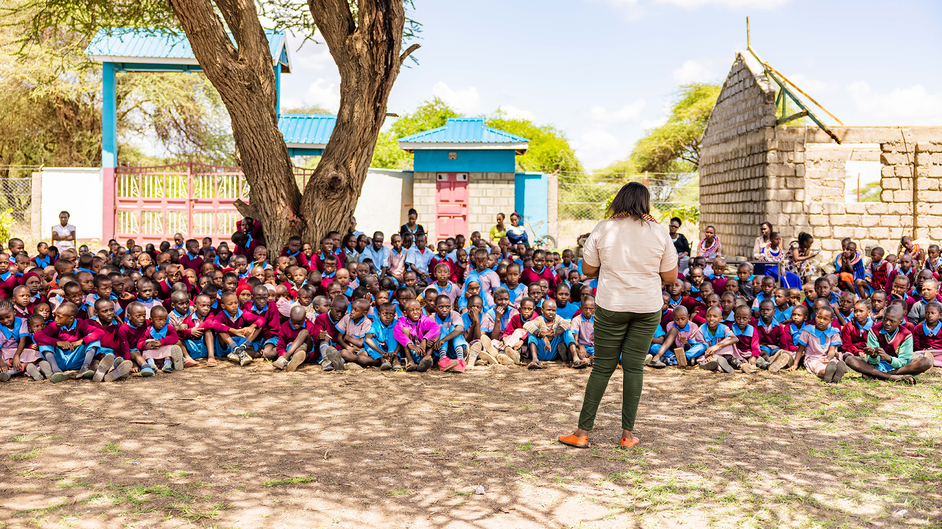 A woman stands with her back to the camera, facing a crowd of dozens of Kenyan school children sitting on the ground