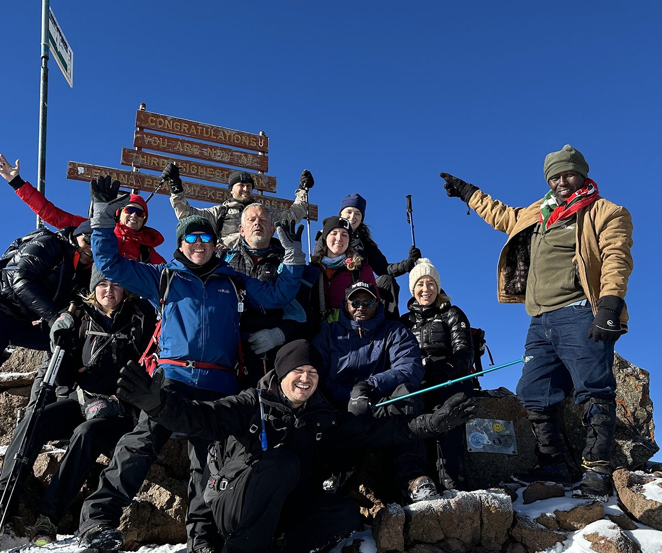 A group of people in cold weather hiking clothes pointing at a Mount Kenya sign against clear blue sky