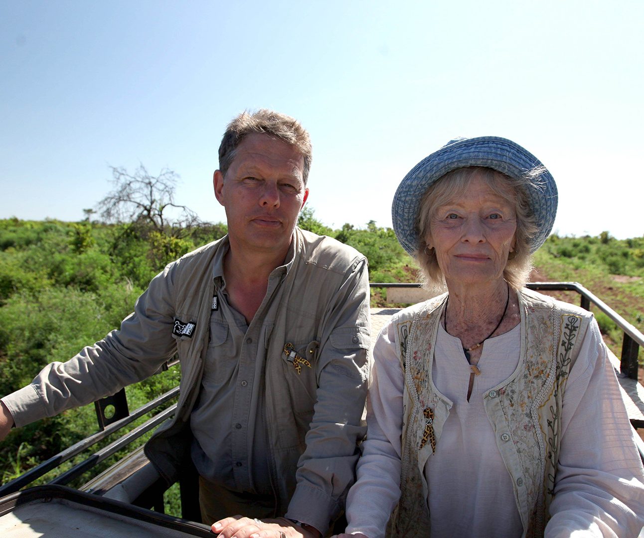 Will Travers and Virginia McKenna are stood side by side in the back of a truck in bright sunlight