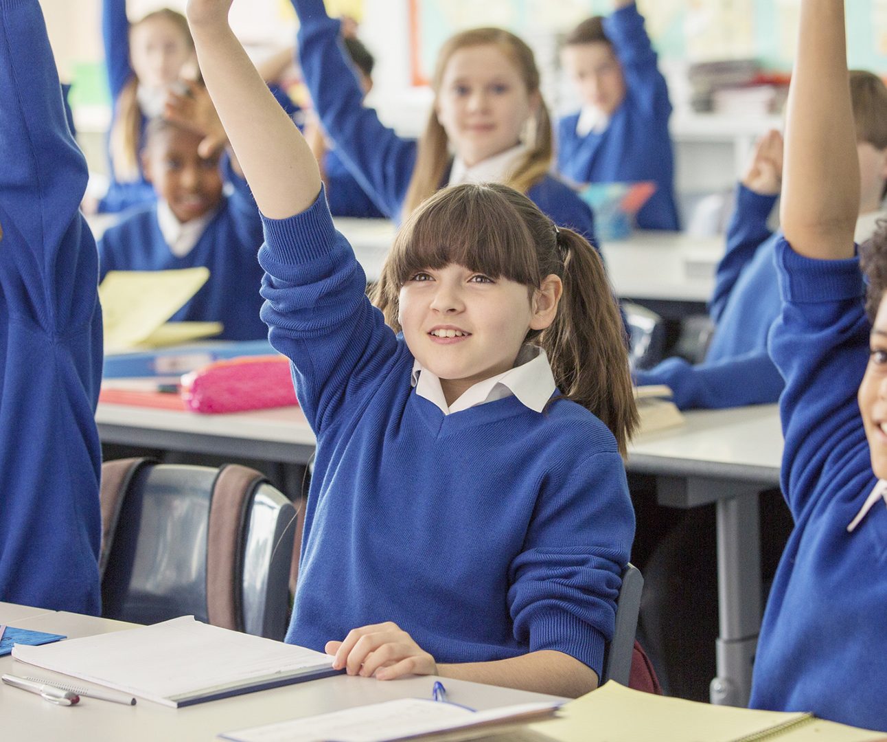 School children in blue jumpers sitting with their hands raised