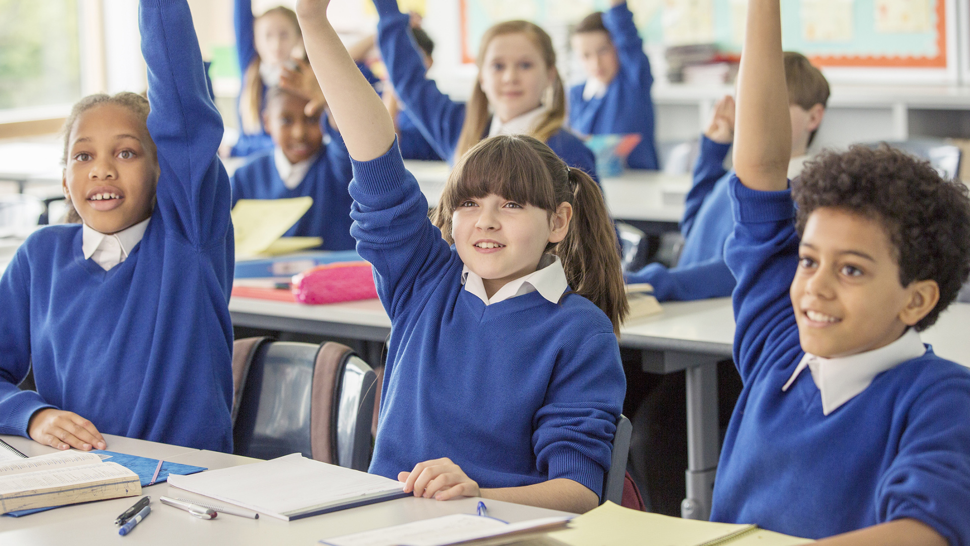 School children in blue jumpers sitting with their hands raised