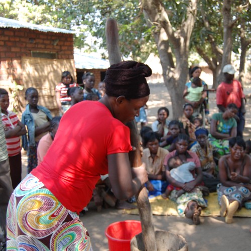 A photo of a village scene in Zambia - a woman prepares food surrounded by children and community members