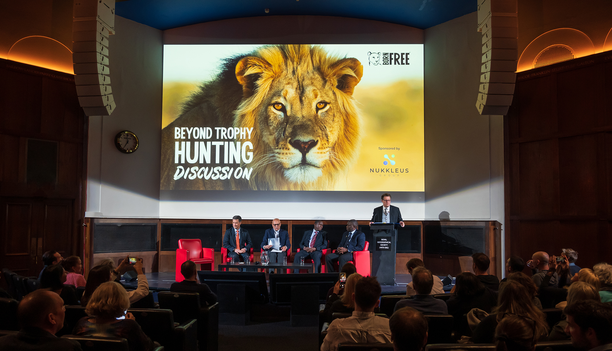 Will Travers standing at a lectern with four panellists at the RGS in London, with 'Beyond Trophy Hunting' and a photo of a lion on the screen behind him