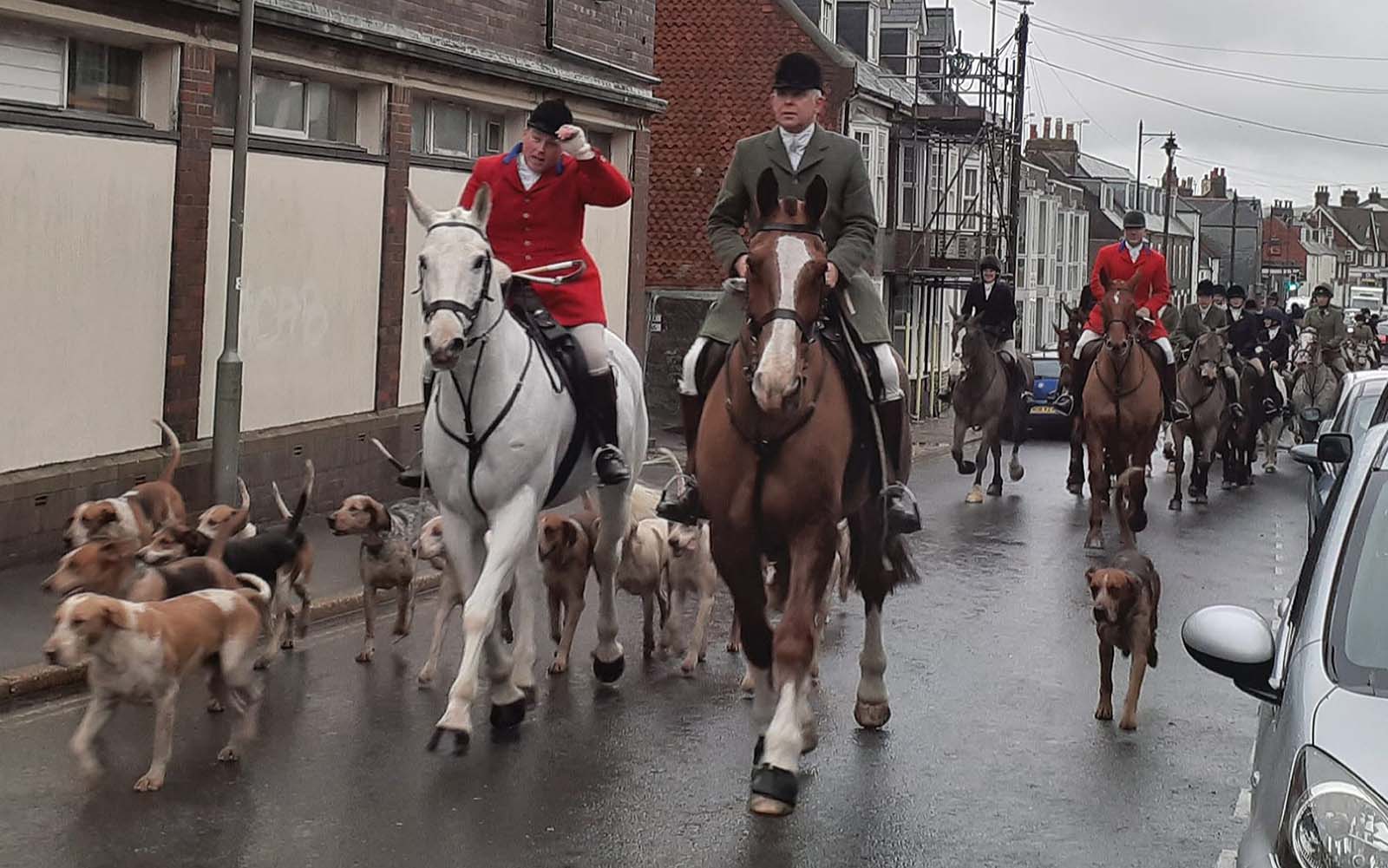 A picture of a boxing day trail hut going through a street, there are numerous people on horseback and hounds.