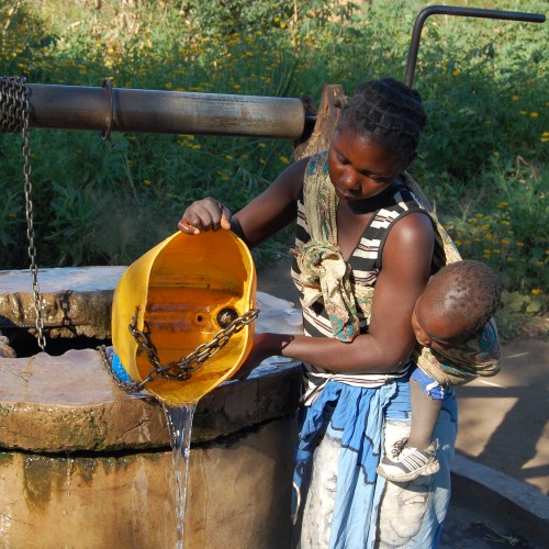 A woman with a young baby strapped to her back gathers water from a well