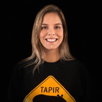 A portrait of a white women with shoulder length blonde hair, she is wearing a black t-shirt which an orange road sign on which says 'Tapir' and standing against a black background.