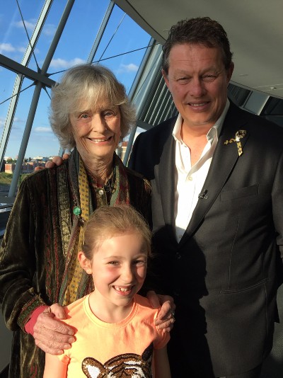 Virginia McKenna stands on the left, with Will Travers on the right, with nine year old Thea Caine between them. Blue sky in the background and everyone  is smiling!