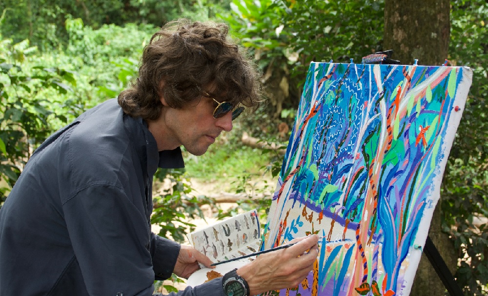 Artist, John Dyer, stands in the rainforest, painting the scene around him. His canvas is covered in colourful plants and trees. 
