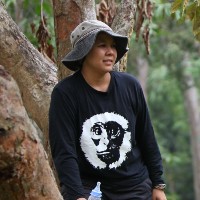 A photo of an Asian woman leaning against a tree in the forest, she is wearing a black longsleeve top, black trousers and a khaki hat. She is holding a bottle of milk in her right hand.