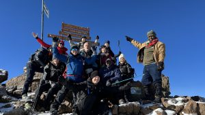 A group of people in cold weather hiking clothes pointing at a Mount Kenya sign against clear blue sky