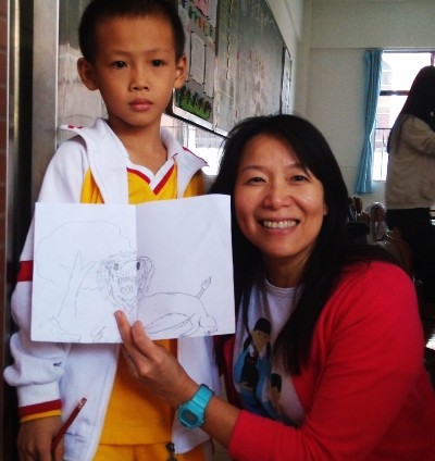 A photo of an Asian woman and an Asian boy in a classroom, the woman is crouched down next to the boy and holding a drawing of a lion and a tree.