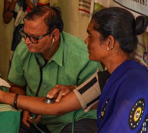 A woman in a blue traditional Indian dress sits side-on to the camera, while she has her blood pressure taken by a man in a green shirt with a stethoscope around his neck.