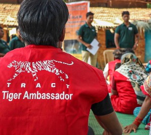 A man with a red 'tiger ambassador' shirt sits with his back to the camera, whilst men in green shirts stand in front of an audience of local people.