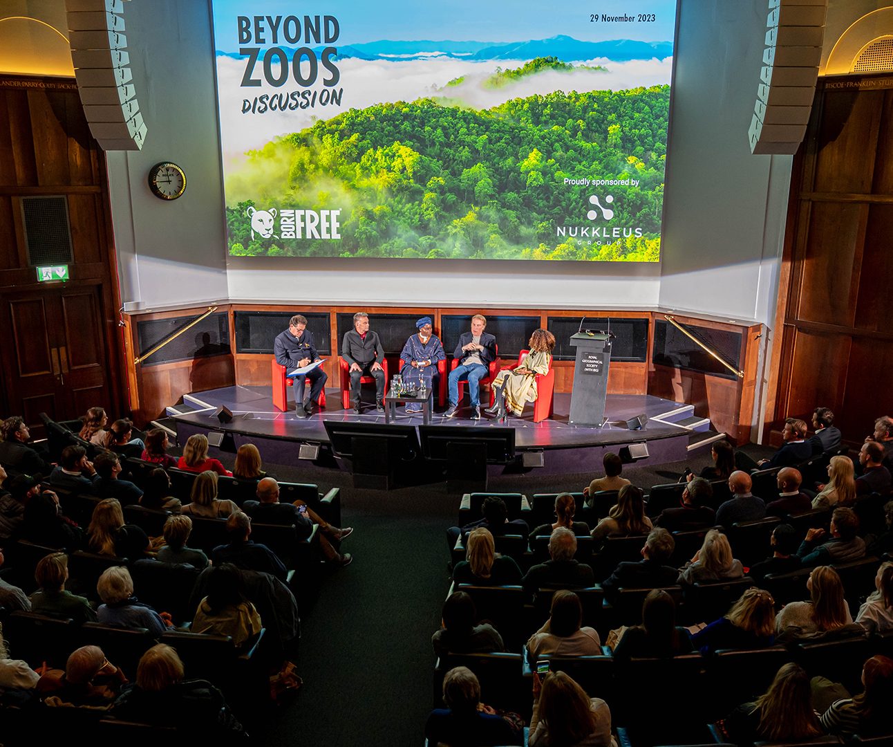 A panel of five people sitting on a stage, with large projector screen behind them and large audience in front