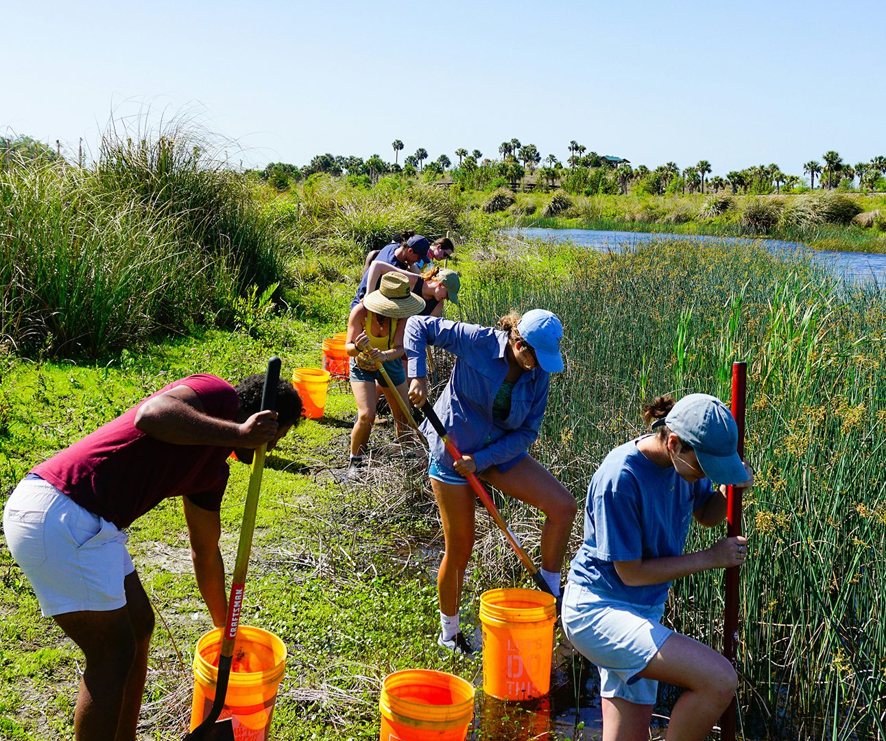 A group of people with orange buckets are lined up along the edge of a river bank, digging with shovels