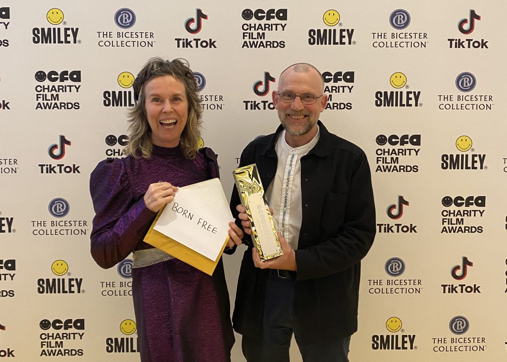 Celia Nicholls and Dr Mark Jones holding an envelope and gold award in front of a step and repeat board
