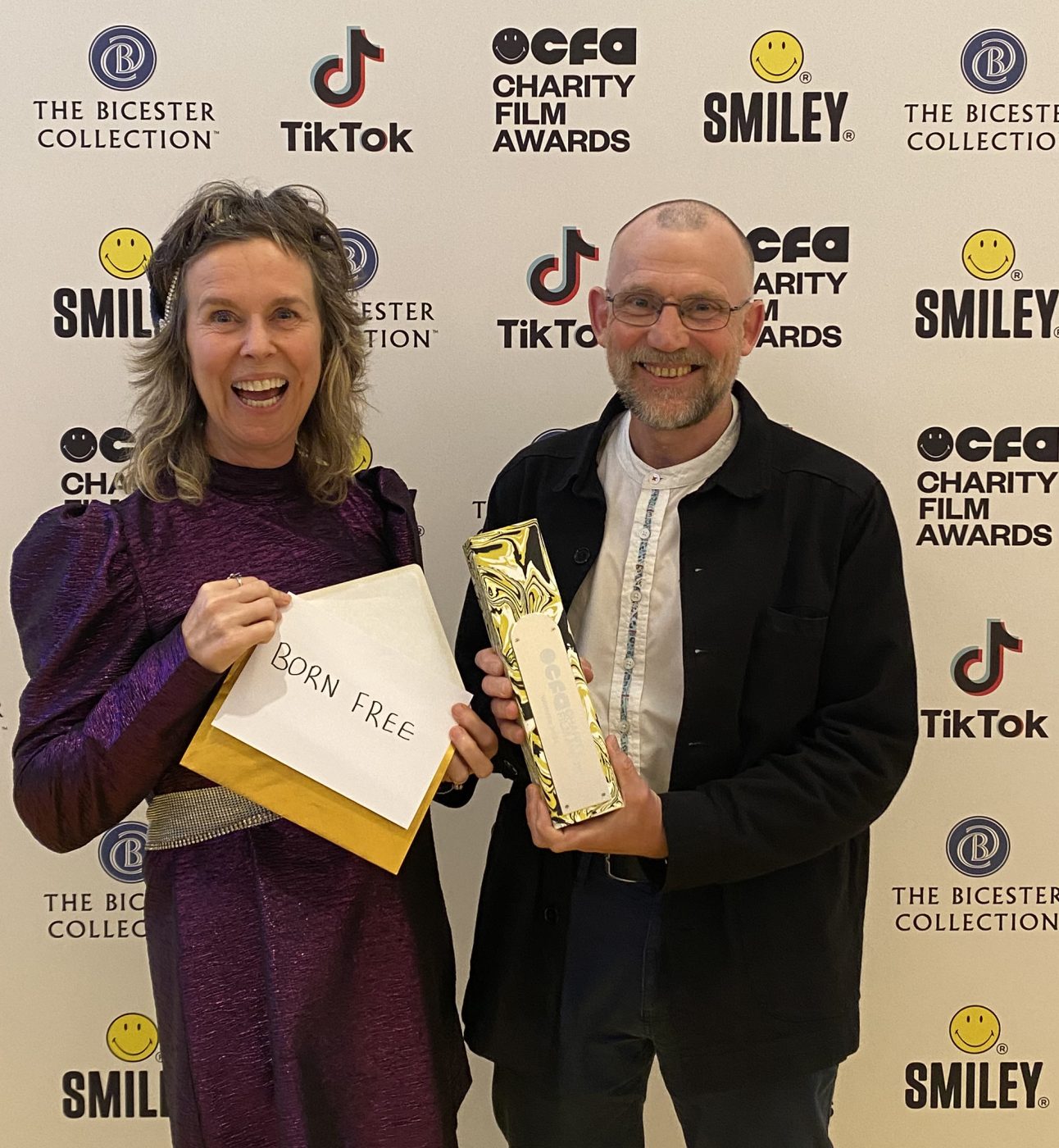 Celia Nicholls and Dr Mark Jones holding an envelope and gold award in front of a step and repeat board