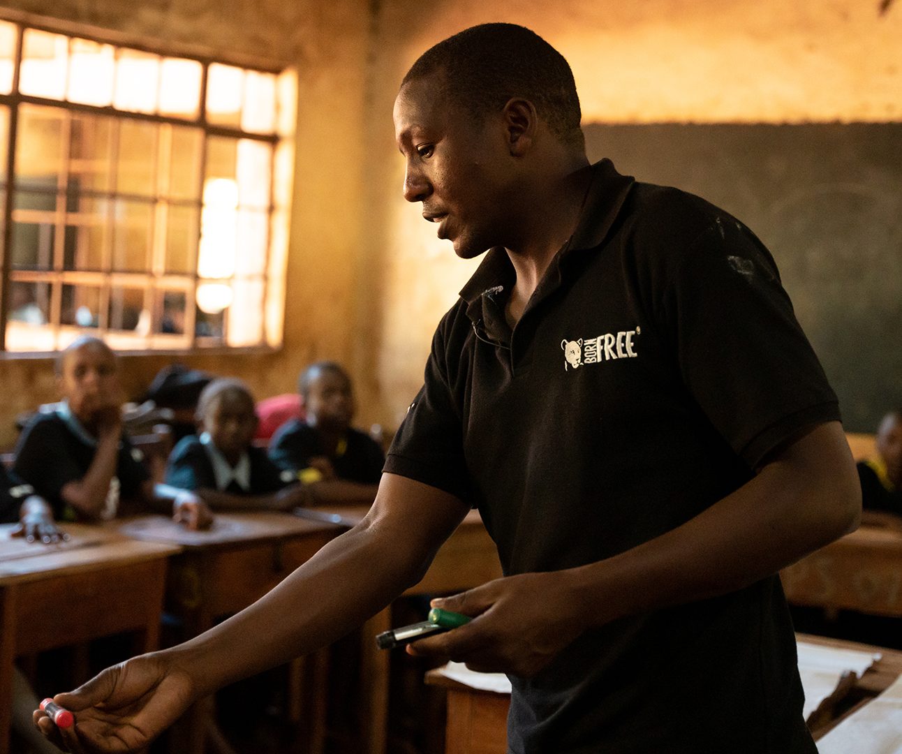 A Kenyan classroom with children in the background and a man in Born Free shirt at the front