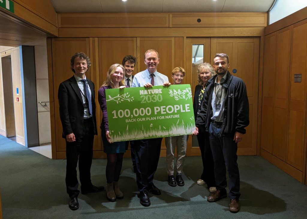 A group of people in an office, holding a large sign saying 'Nature 2030: 100,000 people back our plan for nature'