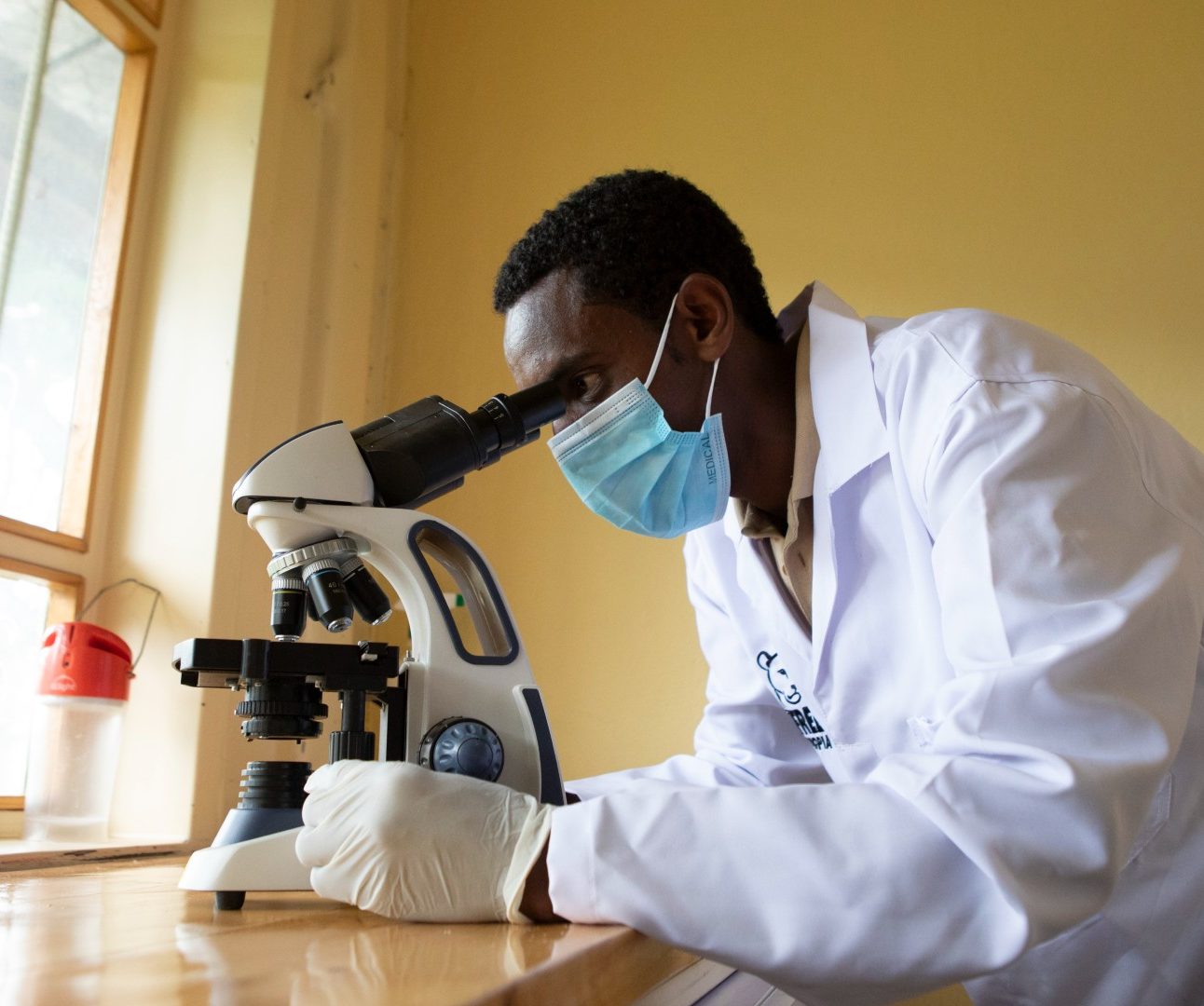 A man wearing a white lab coat and face mask, looking at samples through a microscope