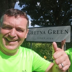 A man wearing a green T-shirt giving a thumbs up as he stands next to a road sign for Gretna Green