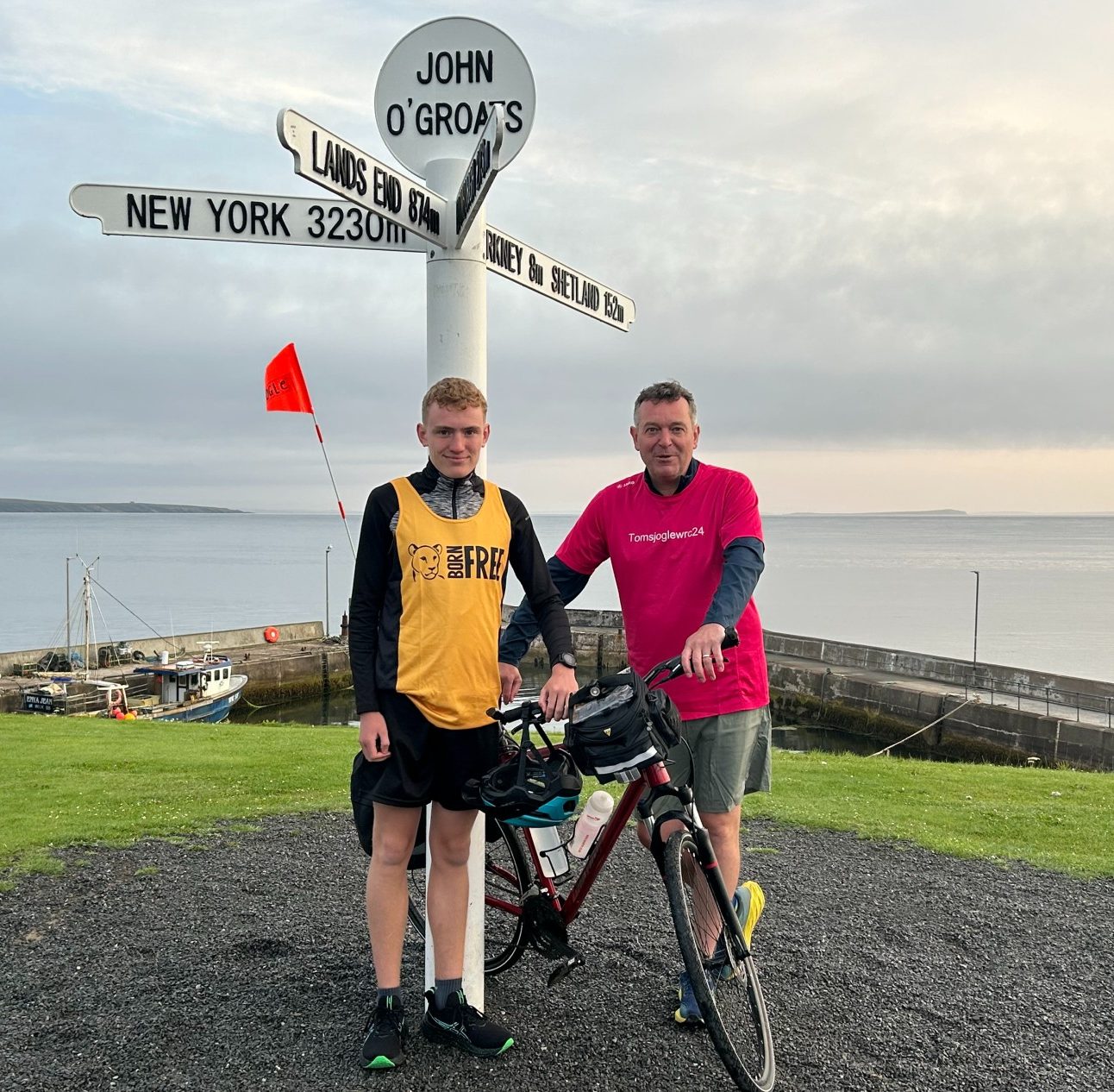 A young man wearing a Born Free running vest standing with his dad at the John O'Groats sign post