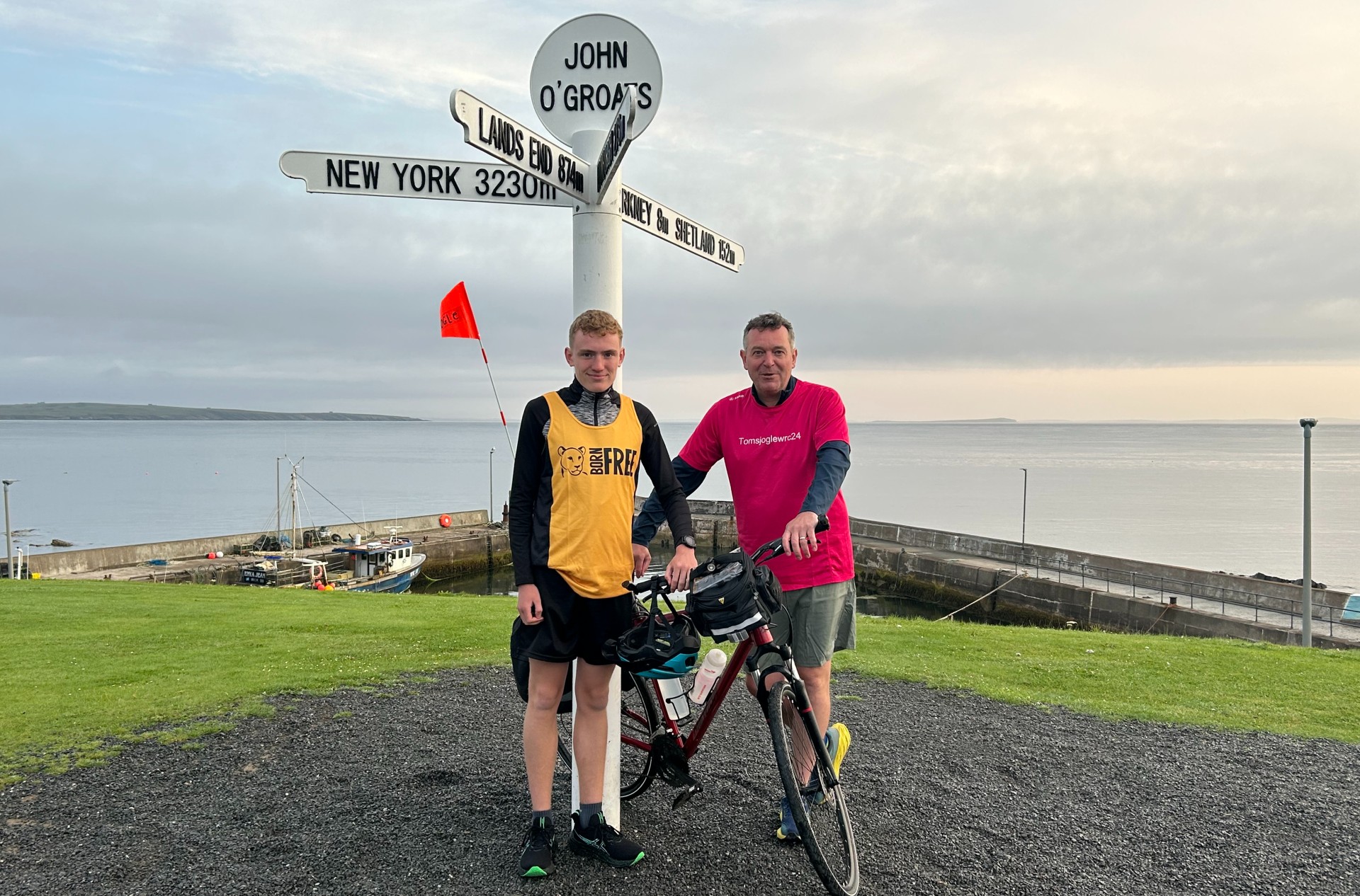 A young man wearing a Born Free running vest standing with his dad at the John O'Groats sign post