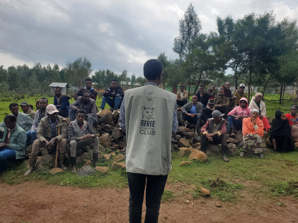 A man in Born Free Ethiopia shirt has his back to the camera, speaking to a large group of people sat outside