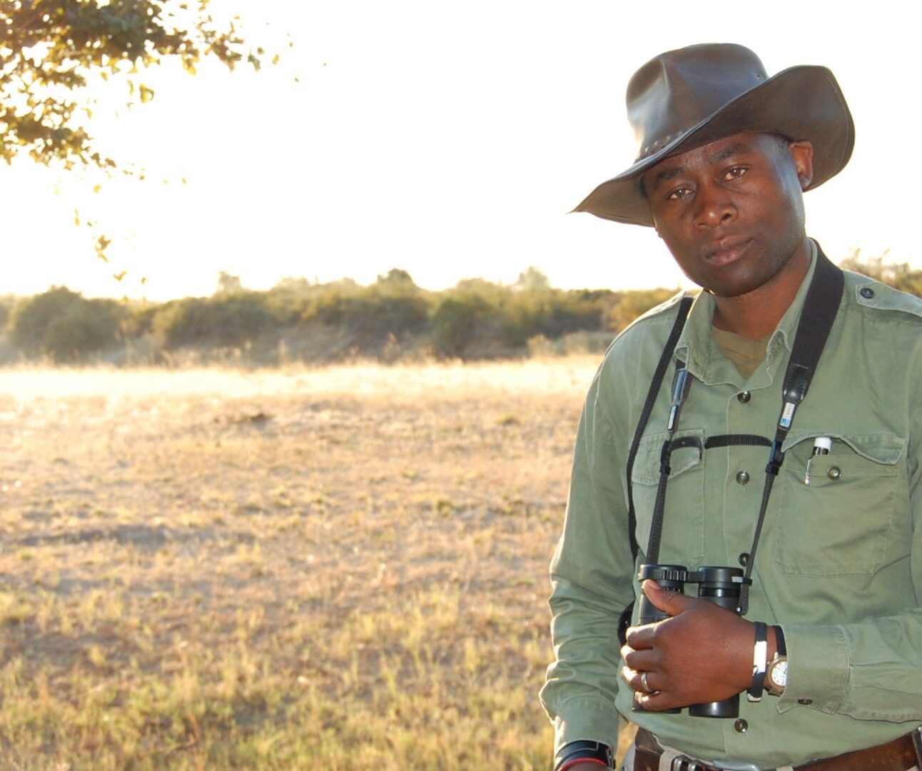 A photo of Manny Mvula standing in the open countryside, holding binoculars