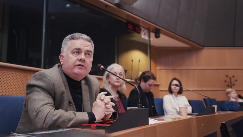 Dominic Dyer is sitting at a desk with a microphone in front of him, with a row of people in the background