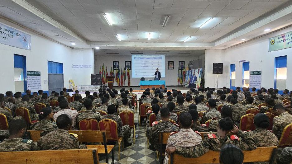 A room is filled with people in military uniform, seated in rows, with a presentation at the front of the room.