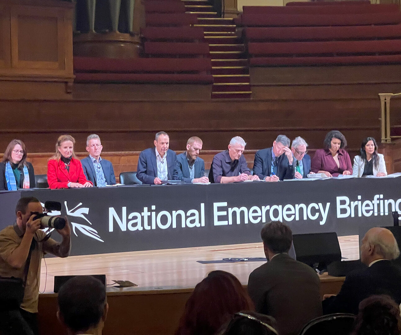 A panel of people sit behind a long desk on a stage, a banner reads National Emergency Briefing