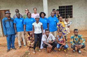 Photograph of a group of 14 people standing and sitting outside a building with a concrete wall and barred window. Several individuals wear matching blue shirts, while others wear colorful patterned clothing, suggesting a community or organizational gathering.