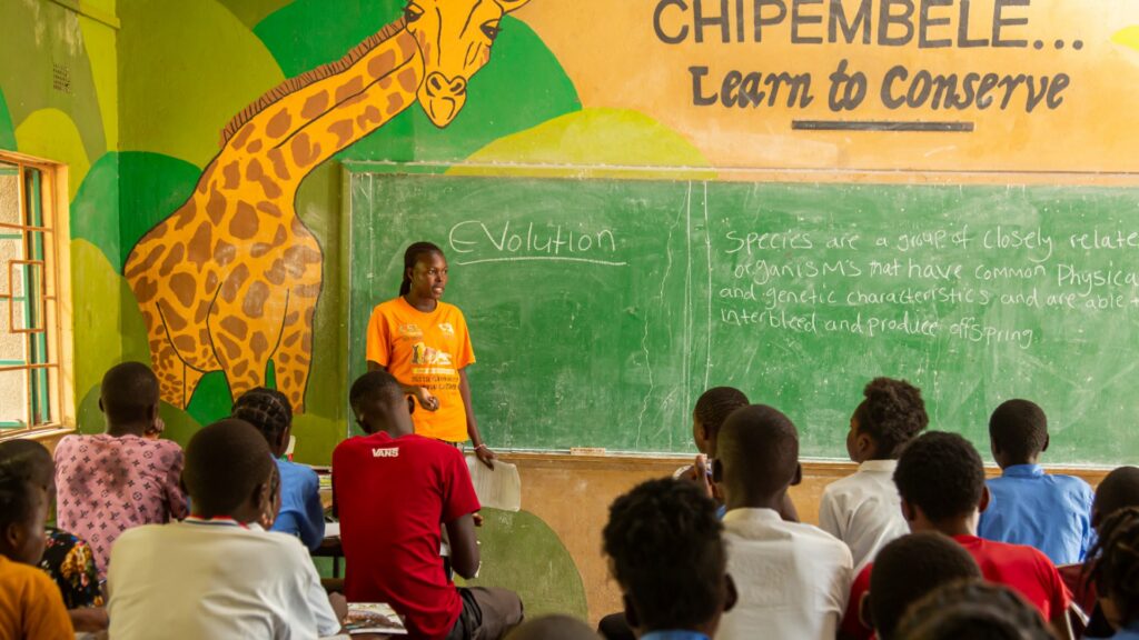 Photograph of a classroom setting with a teacher standing in front of a chalkboard, explaining a lesson on evolution to seated students. The classroom features a large painted giraffe and the phrase "CHIPEMBELE... Learn to Conserve" on the wall, emphasizing conservation education.
