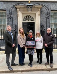Photograph of five people standing in front of a black door with number 10, holding a box labeled "Time for Change." The setting suggests a formal or political context, with individuals dressed in business and casual attire, highlighting a campaign or advocacy event.