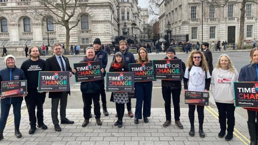 Photograph of a group of people standing outdoors in front of a historic building, holding signs that read "TIME FOR CHANGE" in bold letters with colorful accents. The scene suggests a peaceful demonstration or awareness campaign focused on advocating for change, with participants dressed in casual and professional attire.