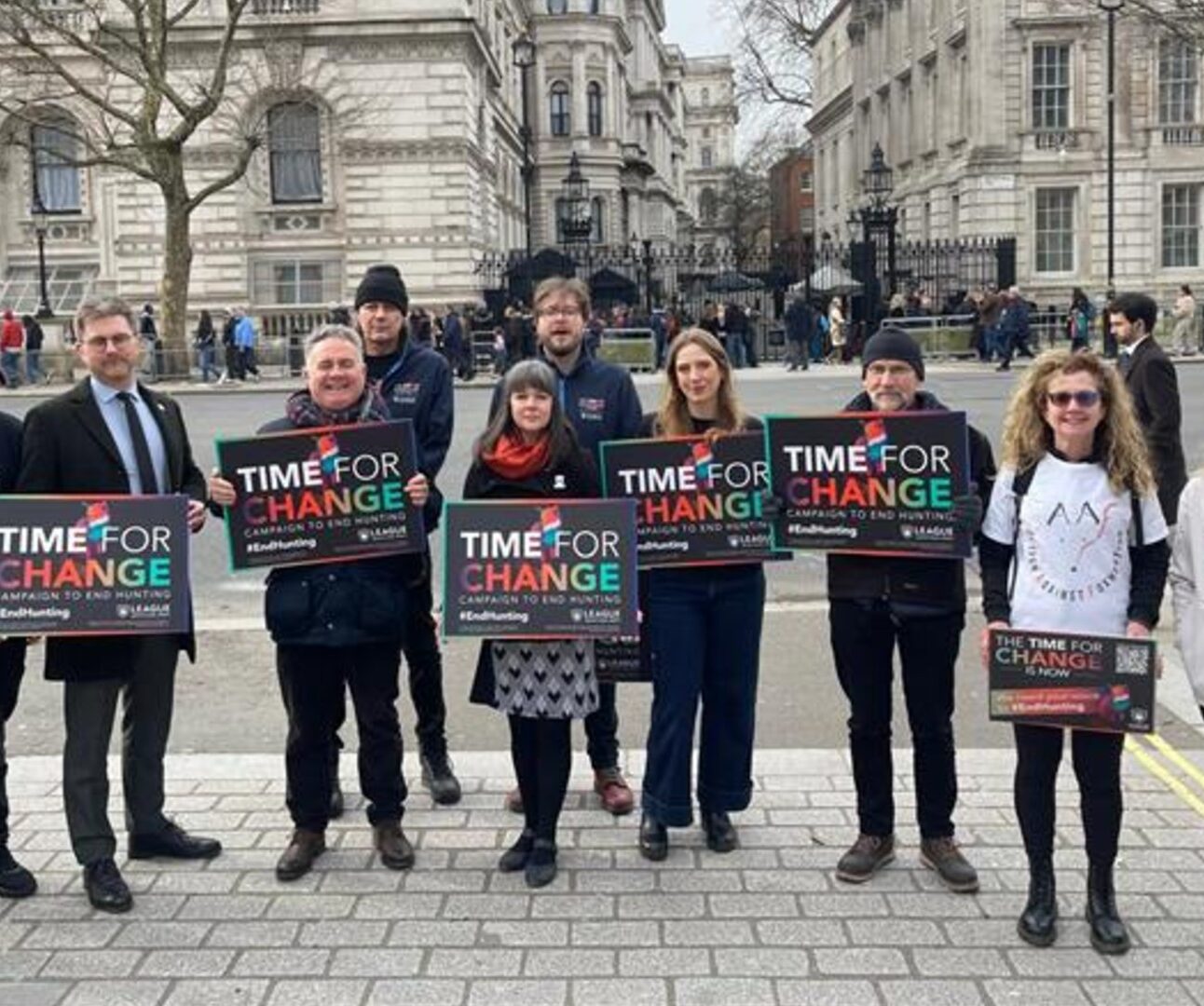 Photograph of a group of people standing outdoors in front of a historic building, holding signs that read 