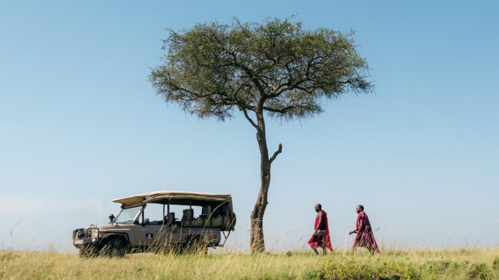 Photograph of a safari vehicle parked under a solitary tree in a grassy savanna with two people dressed in traditional attire walking nearby. The scene captures a peaceful moment in a natural landscape with clear blue sky and tall grass.