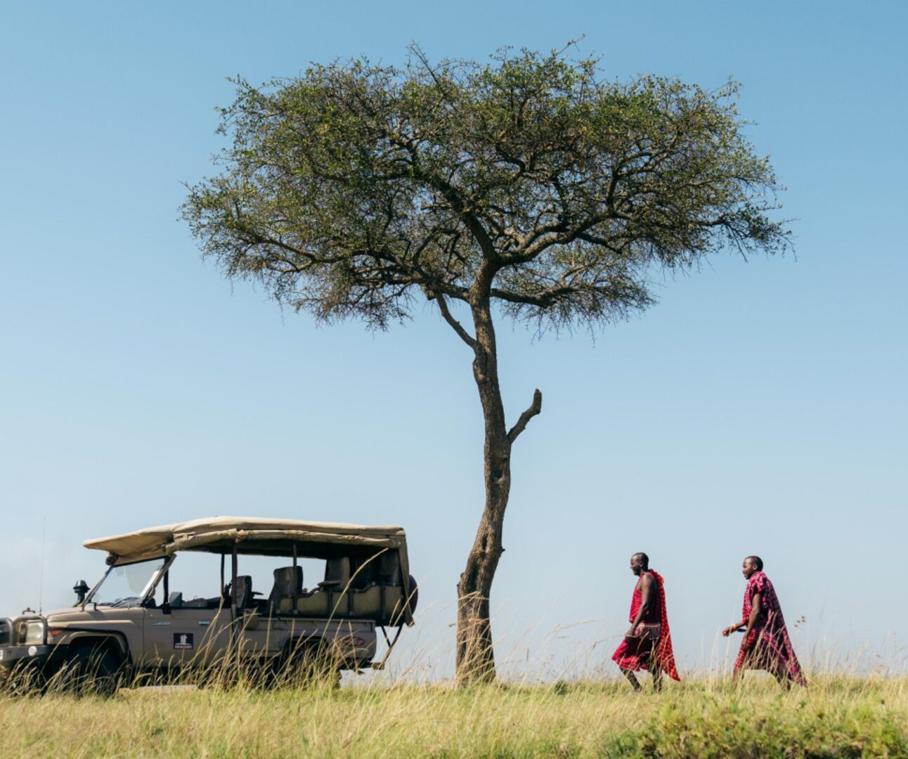 Photograph of a safari vehicle parked under a solitary tree in a grassy savanna with two people dressed in traditional attire walking nearby. The scene captures a peaceful moment in a natural landscape with clear blue sky and tall grass.
