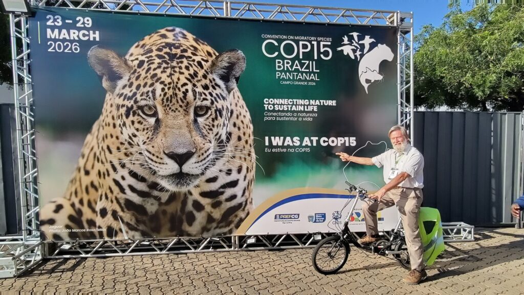 Photograph of a man posing with a bicycle in front of a large promotional banner for COP15 Brazil Pantanal, scheduled for March 23-29, 2026. Banner features a close-up of a jaguar, event logo, tagline "Connecting Nature to Sustain Life," and text "I Was at COP15" in English and Portuguese.
